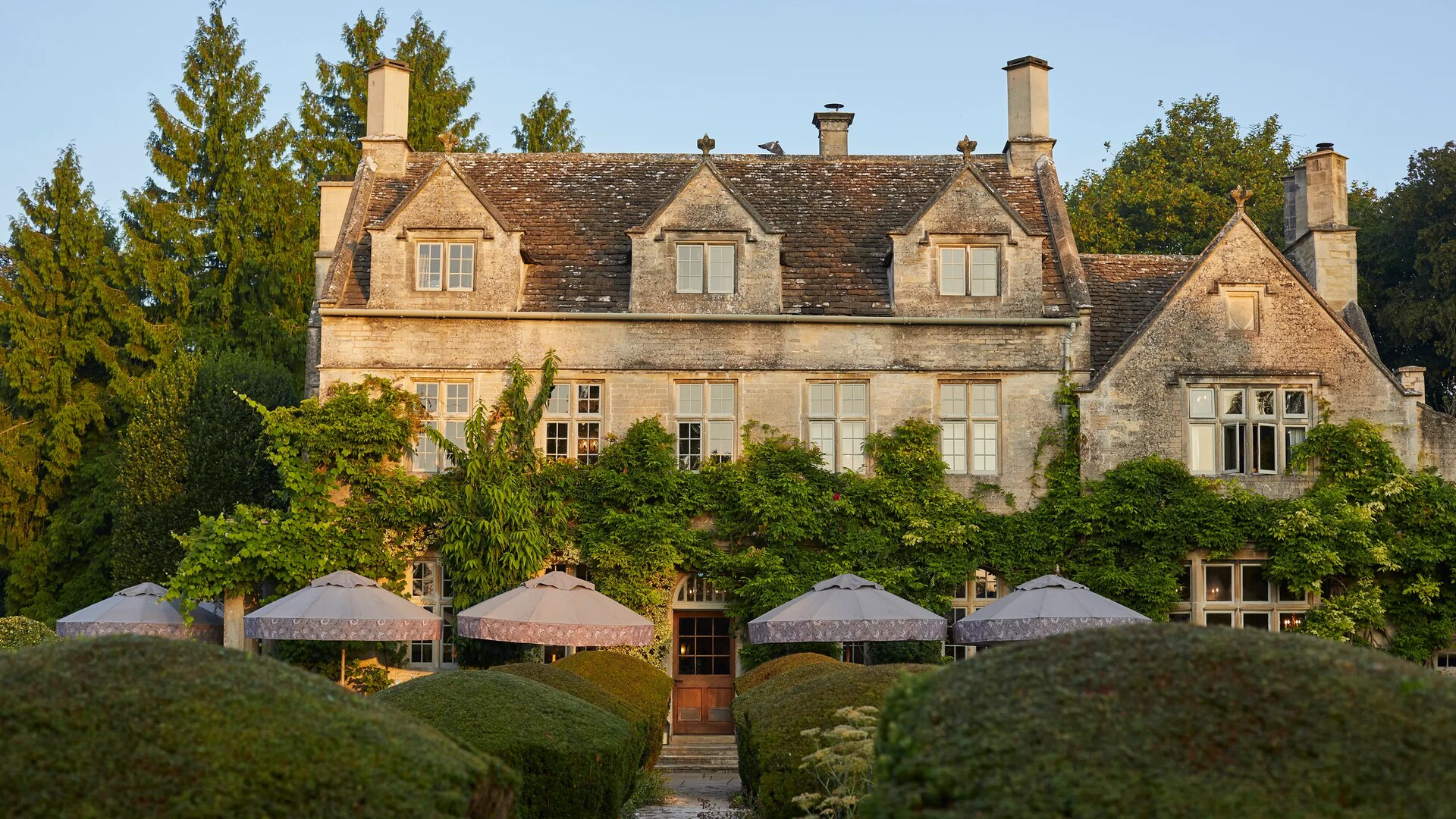 The Pig in the Cotswolds — the honey-stone manor at dusk, with parasols on the lawn